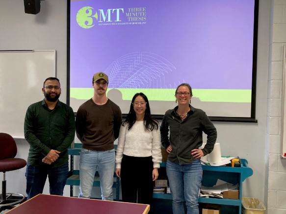 Four students dressed in casual wear stand against a backdrop of a three minute thesis slide.